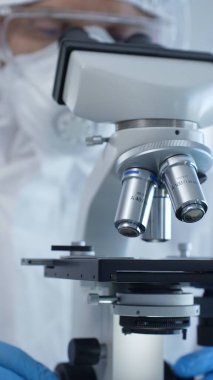 Tehlikeli madde giysisi, maskesi ve eldivenleri olan bir tıp virologu laboratuvar araştırmalarında mikroskop başında özenle çalışıyor. Tıp ve bilim konsepti.