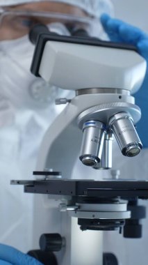 Tehlikeli madde giysisi, maskesi ve eldivenleri olan bir tıp virologu laboratuvar araştırmalarında mikroskop başında özenle çalışıyor. Tıp ve bilim konsepti.
