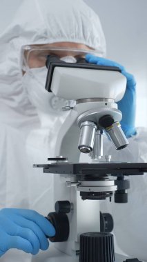 Tehlikeli madde giysisi, maske ve mavi eldiven giyen erkek bir virüs bilimci laboratuvarda virüsleri araştırırken mikroskop altında özenle çalışıyor. Tıp ve bilim konsepti.