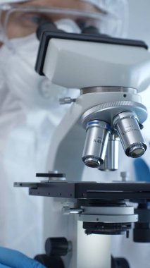 Tehlikeli madde giysisi, maskesi ve eldivenleri olan bir tıp virologu laboratuvar araştırmalarında mikroskop başında özenle çalışıyor. Tıp ve bilim konsepti.