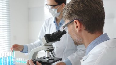 Gözlük takan bir araştırmacı mikroskop altında bir numuneyi inceliyor, koruyucu maske takan bir laboratuvar teknisyeni de çalışma sırasında beyaz laboratuvar önlükleriyle yakınlarda duruyor. Tıp ve Bilim.
