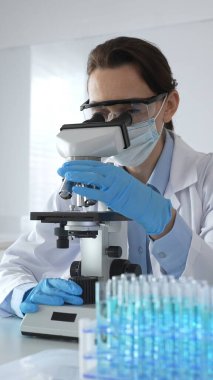 Mikroskop kaydırağını inceleyen kadın bilim adamı, laboratuarda koruyucu giysiler giyiyor, mavi renkli test tüpleriyle farmasötik buluşları araştırıyor..