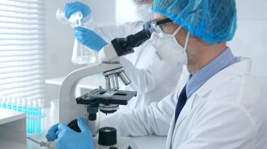 Maskeli erkek bilim adamı mikroskop altında numuneleri incelerken laboratuvar asistanı arka planda profesyonel bir tıbbi araştırmada cam işleriyle uğraşıyor. Tıp, bilim konsepti.