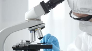 Tehlikeli madde giysisi, maskesi ve mavi eldivenleri olan bir tıp virologu laboratuvarda virüsleri araştırırken mikroskop altında dikkatlice çalışıyor. Tıp ve bilim konsepti.