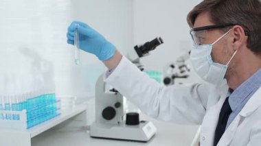 Koruyucu giysi giyen bilim araştırmacısı modern laboratuvar çalışma alanında mavi sıvı numunesini test tüpünden inceliyor. Tıp, sağlık ve bilim konsepti