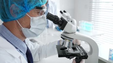 Koruyucu maske, şapka ve eldiven takan kıdemli erkek bilim adamı modern bir laboratuvarda mikroskop altında örnekleri inceliyor. Tıp, sağlık ve bilim konsepti.