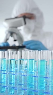 Laboratuvarda koruyucu dişli mikroskop lensleri takan bir bilimsel araştırmacının karşısında mavi sıvıyla test tüplerini kapatın. Tıp, sağlık ve bilim konsepti.
