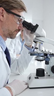 Koruyucu gözlük takan bilim adamı mikroskop altında araştırma ve analiz yapıyor. Arka planda test tüpleri ile modern bir laboratuvarda. Tıp, sağlık ve bilim konsepti