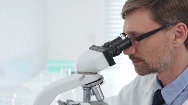 Beyaz önlük ve gözlük giyen erkek bilim adamı mikroskop kullanıyor ve modern bir laboratuvarda bilimsel araştırma yapıyor. Tıp, sağlık ve bilim konsepti.