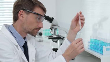 Koruyucu giysi giyen bilim araştırmacısı cam şişeye kimyasal solüsyon döküyor, laboratuvar ekipmanları ve mikroskoplarla çevrili. Tıp, sağlık ve bilim konsepti