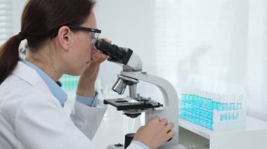 Laboratuvar önlüğü ve koruyucu gözlüklü kadın bilim adamı modern bir laboratuvarda mikroskop altında test örnekleri inceliyor. Tıp, sağlık ve bilim konsepti