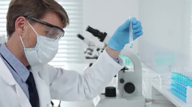 Koruyucu giysi giyen bilim araştırmacısı modern laboratuvar çalışma alanında mavi sıvı numunesini test tüpünden inceliyor. Tıp, sağlık ve bilim konsepti