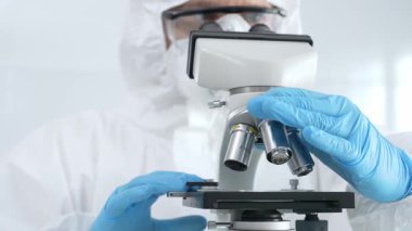 Tehlikeli madde giysisi, maskesi ve mavi eldivenli bir tıp viroloğu bir laboratuvar araştırma virüsünde mikroskop altında dikkatlice çalışıyor. Sağlık, tıp ve bilim konsepti.