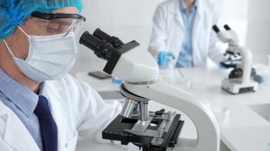 Koruyucu maske, şapka ve eldiven takan kıdemli erkek bilim adamı modern bir laboratuvarda mikroskop altında örnekleri inceliyor. Tıp, sağlık ve bilim konsepti.