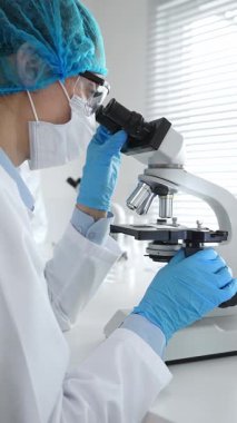 Maske takan ve mavi güvenlik donanımı takan kadın bir bilim adamı araştırma için mikroskop kullanırken erkek bir meslektaşı arka planda çalışıyor. Tıp, sağlık ve bilim konsepti.