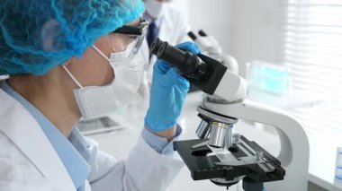 Maske takan ve mavi güvenlik donanımı takan kadın bir bilim adamı araştırma için mikroskop kullanırken erkek bir meslektaşı arka planda çalışıyor. Tıp, sağlık ve bilim konsepti.