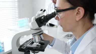 Koruyucu gözlük takan kadın bilim adamı mikroskop kullanarak modern laboratuvarda deney tüpleri ve arka planda erkek bilim adamı ile araştırma yapıyor. Tıp, sağlık ve bilim konsepti