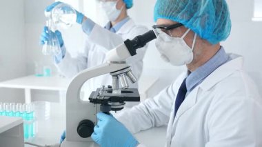 Koruyucu maske takan ve gözlük takan erkek bilim adamı mikroskop kullanarak laboratuvarda araştırma yapıyor ve elinde mataralar olan bir bilim adamı asistanı var. Sağlık ve bilim konsepti.