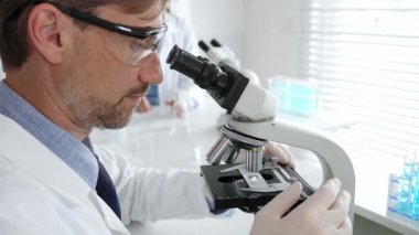 Koruyucu gözlük takan bilim adamı mikroskop altında araştırma ve analiz yapıyor. Arka planda test tüpleri ile modern bir laboratuvarda. Tıp, sağlık ve bilim konsepti