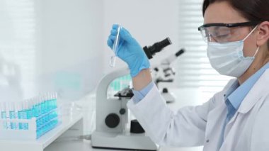 Mavi koruyucu eldivenli maskeli kadın araştırmacı test tüpündeki temiz sıvıyı inceliyor, mikroskop ve laboratuvar ekipmanlarının yanında oturuyor. Tıp, sağlık ve bilim konsepti.