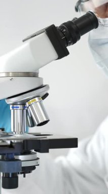 Modern bir laboratuvarda mikroskop altında koruyucu maske, kep ve eldiven takan üst düzey bir bilim adamının örnekleri inceleniyor. Tıp, sağlık ve bilim konsepti.