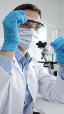 Maske takan ve eldiven takan kadın bilim adamı deney tüpüne sıvı ekliyor. Laboratuvarda tıbbi araştırma için pipet kullanıyor. Tıp, sağlık ve bilim konsepti.