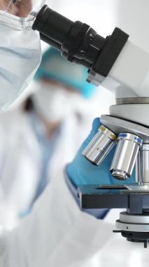 Modern bir laboratuvarda mikroskop altında koruyucu maske, kep ve eldiven takan üst düzey bir bilim adamının örnekleri inceleniyor. Tıp, sağlık ve bilim konsepti.