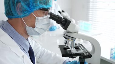 Koruyucu maske, şapka ve eldiven takan kıdemli erkek bilim adamı modern bir laboratuvarda mikroskop altında örnekleri inceliyor. Tıp, sağlık ve bilim konsepti.