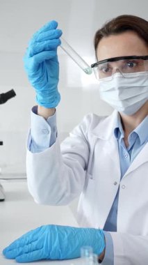 Maske takan, koruyucu gözlük takan ve eldiven takan kadın bilim adamı modern laboratuardaki tıbbi araştırma sırasında mavi sıvıyı dikkatlice inceliyor. Tıp, sağlık ve bilim konsepti