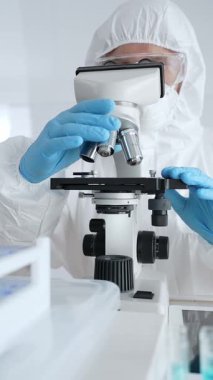 Koruyucu ekipman takan bilim araştırmacısı mikroskobu ayarlıyor, laboratuvar örneklerini steril araştırma ortamında hassasiyetle analiz ediyor. Tıp, sağlık ve bilim konsepti.