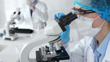 Maske takan ve mavi güvenlik donanımı takan kadın bir bilim adamı araştırma için mikroskop kullanırken erkek bir meslektaşı arka planda çalışıyor. Tıp, sağlık ve bilim konsepti.