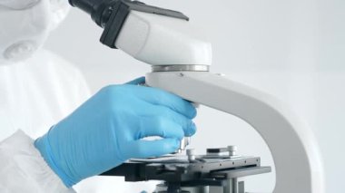 Tehlikeli madde giysisi, maskesi ve mavi eldivenleri olan bir tıp virologu laboratuvarda virüsleri araştırırken mikroskop altında dikkatlice çalışıyor. Tıp ve bilim konsepti.