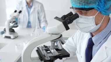 Koruyucu maske, şapka ve eldiven takan kıdemli erkek bilim adamı modern bir laboratuvarda mikroskop altında örnekleri inceliyor. Tıp, sağlık ve bilim konsepti.