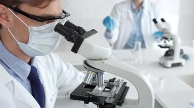 Mikroskop kullanarak maske takan ve eldiven takan bilim adamlarına yakın çekim, modern laboratuvarda araştırmalar yürütmek ve arka planda çalışmak. Tıp, sağlık ve bilim konsepti