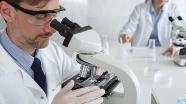 Koruyucu gözlük takan bilim adamı mikroskop altında araştırma ve analiz yapıyor. Arka planda test tüpleri ile modern bir laboratuvarda. Tıp, sağlık ve bilim konsepti