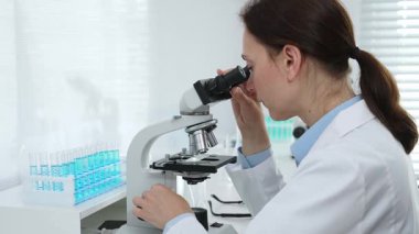 Laboratuvar önlüğü ve koruyucu gözlüklü kadın bilim adamı modern bir laboratuvarda mikroskop altında test örnekleri inceliyor. Tıp, sağlık ve bilim konsepti