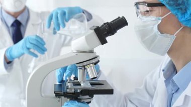 Koruyucu giysili kadın bilim adamı mikroskop altında numuneleri incelerken laboratuvar asistanı profesyonel bir tıbbi araştırmada cam ile ilgileniyor. Tıp ve bilim konsepti.
