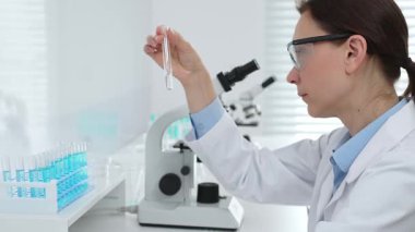 Laboratuvar önlüğü giyen ve koruyucu gözlük takan kadın bilim adamı modern laboratuarda mikroskop ve test tüpleri askısı ile test tüpünü temiz sıvıyla analiz ediyor. Tıp, sağlık ve bilim konsepti