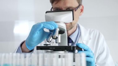 Beyaz önlüklü ve mavi eldivenli erkek bilim adamı mikroskobu ayarlıyor, modern laboratuarda ilaç araştırması yapıyor. Tıp, sağlık ve bilim konsepti.