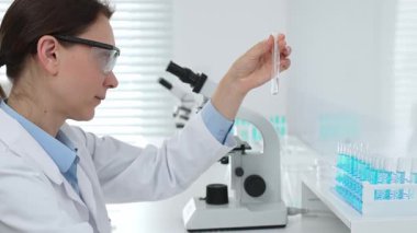 Laboratuvar önlüğü giyen ve koruyucu gözlük takan kadın bilim adamı modern laboratuarda mikroskop ve test tüpleri askısı ile test tüpünü temiz sıvıyla analiz ediyor. Tıp, sağlık ve bilim konsepti