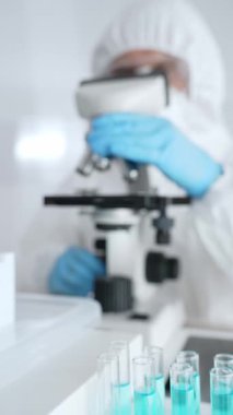 Laboratuvarda koruyucu dişli mikroskop lensleri takan bir bilimsel araştırmacının karşısında mavi sıvıyla test tüplerini kapatın. Tıp, sağlık ve bilim konsepti.