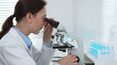 Laboratuvar önlüğü ve koruyucu gözlüklü kadın bilim adamı modern bir laboratuvarda mikroskop altında test örnekleri inceliyor. Tıp, sağlık ve bilim konsepti