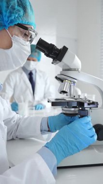 Maske takan ve mavi güvenlik donanımı takan kadın bir bilim adamı araştırma için mikroskop kullanırken erkek bir meslektaşı arka planda çalışıyor. Tıp, sağlık ve bilim konsepti.