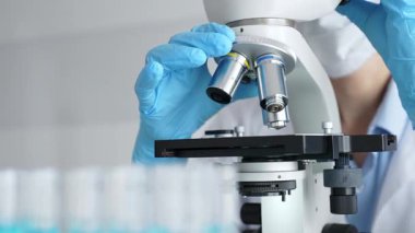 Mikroskobu ayarlayan eldivenli bilim adamlarına yakın çekim yapmak, laboratuvarda araştırma yapmak ve arka planda mavi sıvı içeren test tüpleri kullanmak. Tıp, sağlık ve bilim konsepti.
