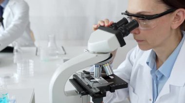 Koruyucu gözlük takan kadın bilim adamı mikroskop kullanarak modern laboratuvarda deney tüpleri ve arka planda erkek bilim adamı ile araştırma yapıyor. Tıp, sağlık ve bilim konsepti