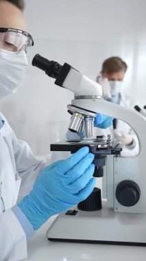 Kadın araştırmacı mikroskop ile örnek analiz ediyor, koruyucu teçhizat takılı, steril araştırma ortamında gelişmiş laboratuvar ekipmanlarının yanında çalışan bir meslektaşı. Tıp, sağlık ve bilim konsepti
