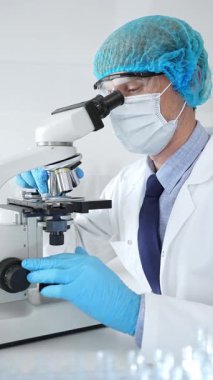 Koruyucu maske, şapka ve eldiven takan kıdemli erkek bilim adamı modern bir laboratuvarda mikroskop altında örnekleri inceliyor. Tıp, sağlık ve bilim konsepti.
