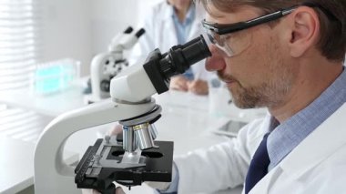 Koruyucu gözlük takan bilim adamı mikroskop altında araştırma ve analiz yapıyor. Arka planda test tüpleri ile modern bir laboratuvarda. Tıp, sağlık ve bilim konsepti