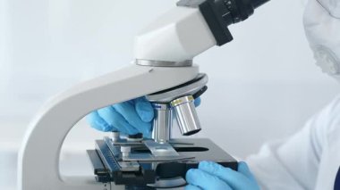 Mavi eldiven giyen bilim araştırmacısı mikroskop altında numuneleri dikkatlice analiz ederek hassas laboratuvar incelemesi yapıyor. Sağlık ve bilim konsepti.