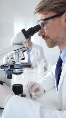 Koruyucu gözlük takan bilim adamı mikroskop altında araştırma ve analiz yapıyor. Arka planda test tüpleri ile modern bir laboratuvarda. Tıp, sağlık ve bilim konsepti